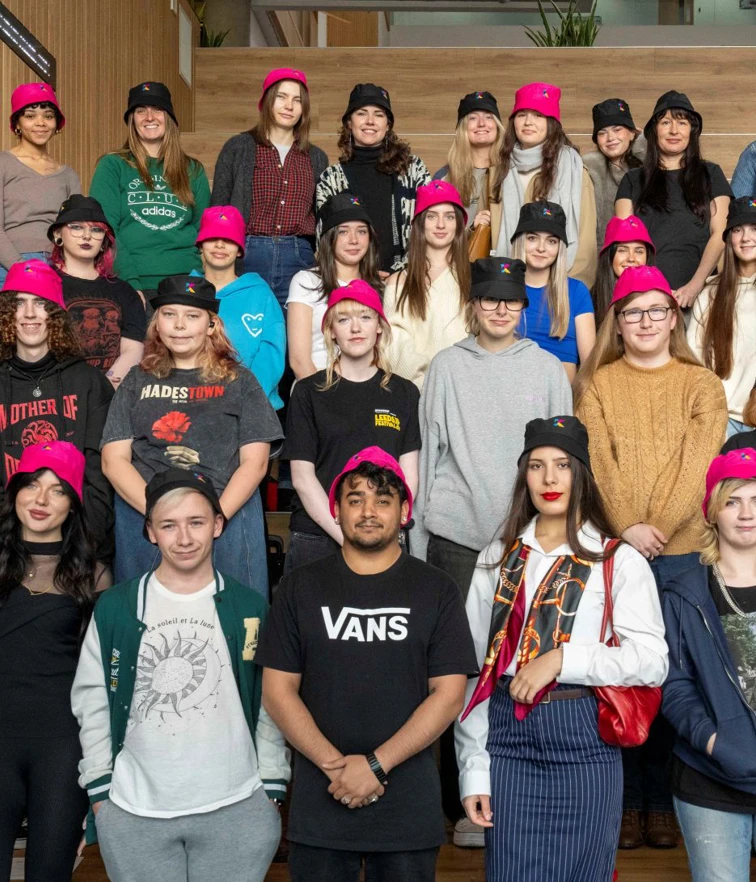 Large group of students wearing pink and black bucket hats, standing on indoor steps at Glasgow Kelvin College, smiling towards the camera Large group of students wearing pink and black bucket hats, standing on indoor steps at Glasgow Kelvin College, smiling towards the camera